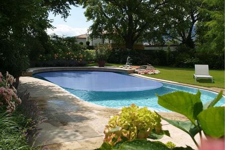 Außenpool mit blauer Abdeckung in einem grünen Garten. Ein weißer Liegestuhl steht am Poolrand.